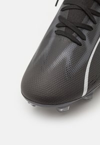 Scarpino da calcio nero con materiale sintetico testurizzato, dotato di lacci neri sottili e un design elegante. Tacchetti trasparenti visibili sulla parte inferiore.