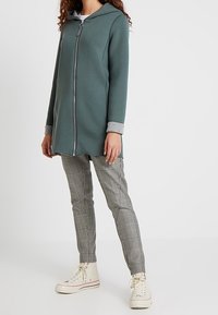 Persoon in een teal kleurige ritssluiting hoodie, grijs geruite slim-fit broek en witte hoge sneakers, staand tegen een effen lichte achtergrond.