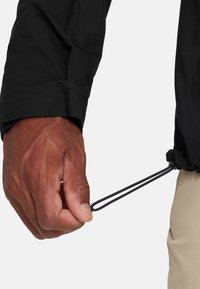 Sort jakke med struktureret stof, der viser en hånd, der griber fat om en snor ved manchetterne, kontrastsyninger og en sikker pasform.
