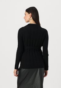 Svart, ribbet cardigan med lange ermer og en tettsittende liv. Har sjalskrage og et knytebelte. Glatt tekstur, enkel design.