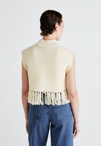 Strikket ermeløs genser i kremfarge, med ribbestrikket krage og frynser langs bunnkanten. Kombinert med høyt liv denimbukser.