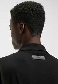 Schwarzes Polohemd mit geripptem Kragen und einem metallischen Logotag auf der Rückseite, das eine glatte Textur und eine taillierte Silhouette präsentiert.