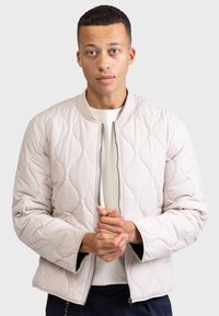 Šviesiai smėlinės spalvos pagalv�ėmis dekoruota bomber tipo striukė su raukšlėtu apykakle, užtrauktuku ir rankogaliais, pasižyminti lygiu paviršiumi ir minkštu užpildu.
