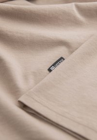 Beige bomullstyg med slät textur, vikt med synlig söm. En liten svart märkesetikett finns i det nedre hörnet.
