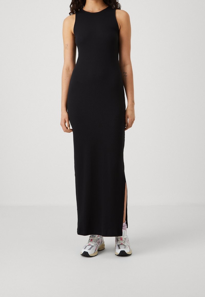Femme portant une robe maxi noire sans manches avec une fente sur le côté et des baskets blanches, debout devant un fond uni.