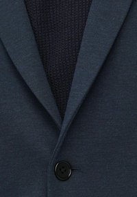 Blazer blu scuro con strato interno a maglia testurizzata, dotato di colletto a becco e chiusura con un singolo bottone nero. Finitura del tessuto liscia e raffinata.