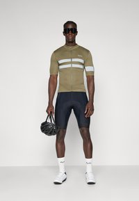 Maillot de cyclisme à manches courtes de couleur vert olive avec des bandes réfléchissantes blanches et une fermeture éclair sur le devant, associé à un short noir et des chaussures de cyclisme blanches.