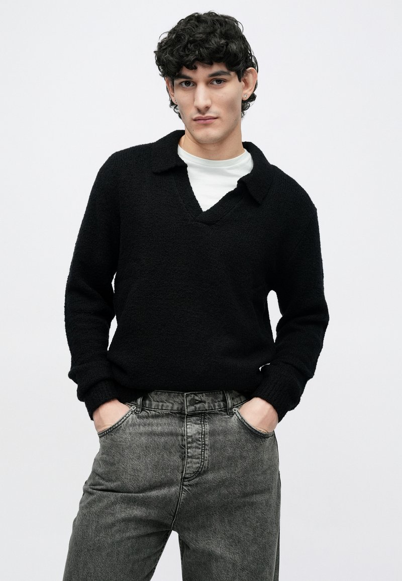 Young man with black curly hair wearing a black sweater over a white shirt and gray jeans, standing with hands in pockets against a plain background.