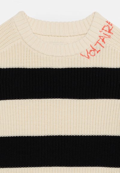 Πλεκτό πουλόβερ με κρεμ και μαύρες ρίγες, με ριμπ λαιμόκοψη και κόκκινη κεντημένη γραφή «Voltaire» κοντά στον ώμο.