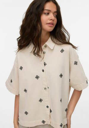 Mujer con cabello largo y oscuro que lleva una camisa de botones de manga corta beige con bordado en forma de rombo negro, mirando hacia su izquierda.