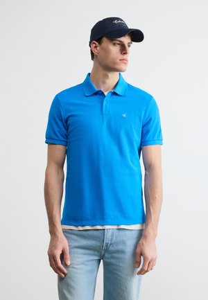 Mladý muž v jasne modrom polokošeli, svetlomodrých džínsoch a tmavomodrej šiltovke, hľadí doľava na jednoduchom pozadí.