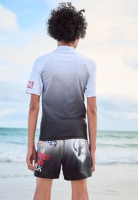 Kortærmet badetrøje med gradient i hvid og grå med et rødt logo, parret med sorte shorts med grafikprint og tekstelementer.