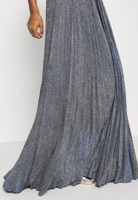 Metallic silver aftonklänning med ett plisserat mönster, med en skimrande yta och blå detaljer, som flödar ner till golvet.