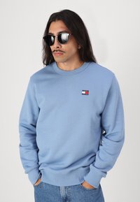 Felpa azzurra con collo rotondo e polsini a costine, presenta un piccolo logo rosso e nero sul lato sinistro superiore. Jeans in denim casual.