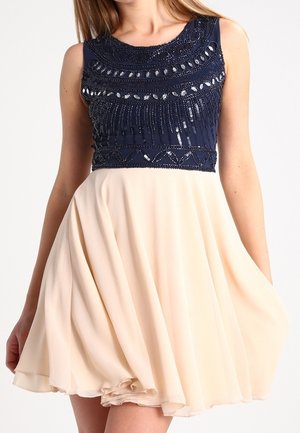 Vrouw draagt een mouwloze jurk met een marineblauwe, met kralen versierde top en een vloeiende beige rok, houdt de rok met één hand lichtjes omhoog.