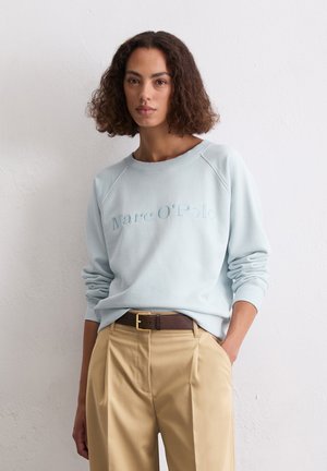 Vrouw met krullend bruin haar in een lichtblauwe Marc O'Polo sweatshirt en beige broek met een bruine riem, staand tegen een witte wand.