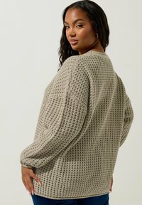 Beige stickad tröja med ett texturerat mönster, rund halsringning, långa ärmar och en avslappnad passform. Har ribbade manschetter och nederkant.