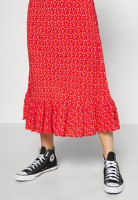 Jambes portant des baskets montantes noires avec des lacets blancs et une jupe midi rouge à fleurs avec un ourlet à volants.