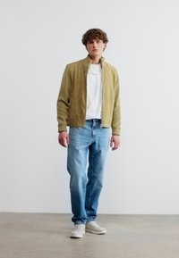 Νεαρός άνδρας που στέκεται φορώντας ελιάς πράσινο μπουφάν με φερμουάρ, λευκό πουκάμισο, ανοιχτό μπλε τζιν και λευκά αθλητικά παπούτσια μπροστά σε απλό λευκό τοίχο.