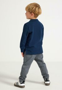 Marine langærmet polo med sidesplids, parret med grå sweatpants. Drengen har sorte og hvide sneakers på og står med ryggen til.
