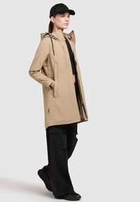Chaqueta impermeable beige con capucha, cremallera frontal y bolsillos laterales, combinada con pantalones cargo de pierna ancha negros y zapatillas. Se lleva una gorra negra.