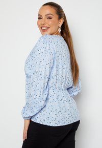 Blusa azzurra chiara con motivo floreale, maniche a sbuffo e dettaglio di vita arricciata. Il modello indossa pantaloni neri e orecchini scintillanti.