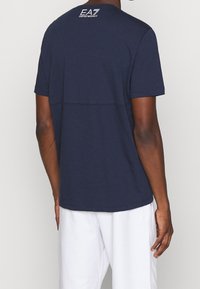 Navy blue short-sleeve t-shirt featuring a crew neck and a horizontal seam detail. Back displays white EA7 logo on the upper neck.