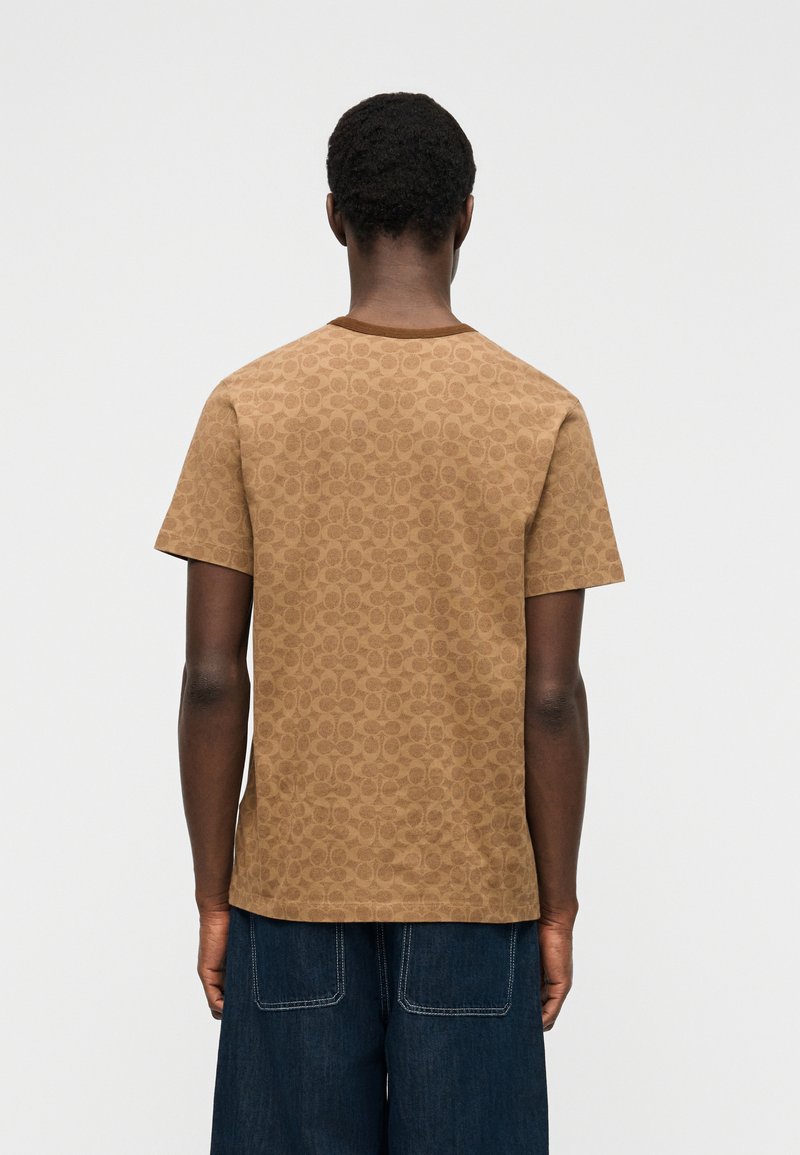 Personne de dos portant une chemise à manches courtes marron à motifs et un pantalon en jean bleu foncé sur fond blanc uni.