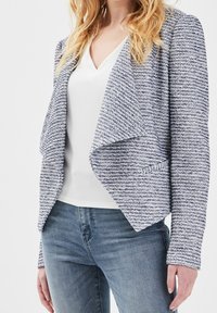 Femme portant un blazer bleu et blanc texturé à ouverture sur le devant, par-dessus un t-shirt blanc à col en V et un jean bleu, debout devant un fond blanc.