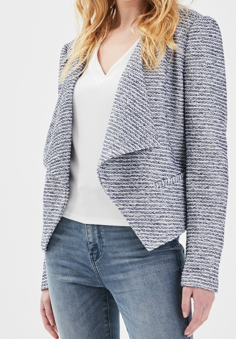 Femme portant un blazer bleu et blanc texturé à ouverture sur le devant, par-dessus un t-shirt blanc à col en V et un jean bleu, debout devant un fond blanc.