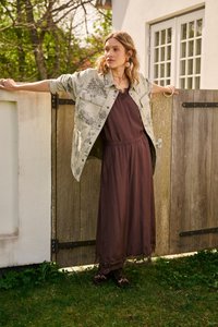 Femme en longue robe marron et veste à motifs, penchée, les bras étendus, sur une porte de jardin en bois dans un cadre extérieur ensoleillé.