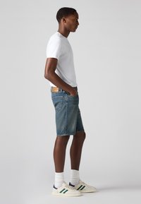 Pantaloni scurți din denim albastru, cu margini ușor sfâșiate, asortați cu un tricou alb și șosete albe. Pantofii sunt bej, cu accente verzi și bleumarin.