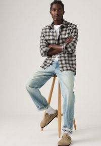 Νέος άνδρας με πλεξούδες κάθεται σε ξύλινο σκαμπό, φοράει ασπρόμαυρο καρό πουκάμισο, λευκό μπλουζάκι, ανοιχτό μπλε τζιν, λευκές κάλτσες και μπεζ παντόφλες.