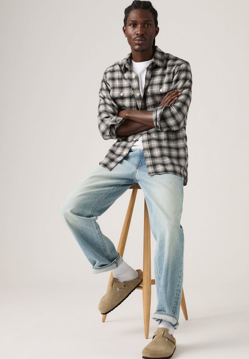 Jeune homme aux cheveux tressés assis sur un tabouret en bois, portant une chemise à carreaux noir et blanc, un t-shirt blanc, un jean bleu clair, des chaussettes blanches et des chaussons beiges.