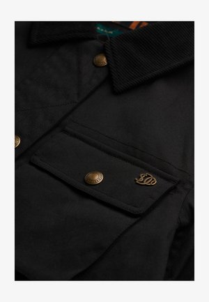 Sort jakke med en tekstureret krave, gyldne dele og en brystlomme med et logo. Materialet virker slidstærkt og glat.