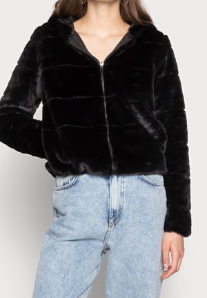 Vrouw draagt een zwarte nepbont jas met rits en lichtblauwe hoge taille jeans, staand met één hand in de zak.
