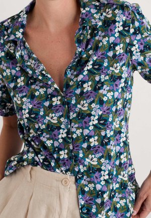 Zbliżenie osoby noszącej zapinaną na guziki, krótką, kwiatową koszulę z fioletowymi, białymi i zielonymi kwiatami oraz beżowe spodnie.