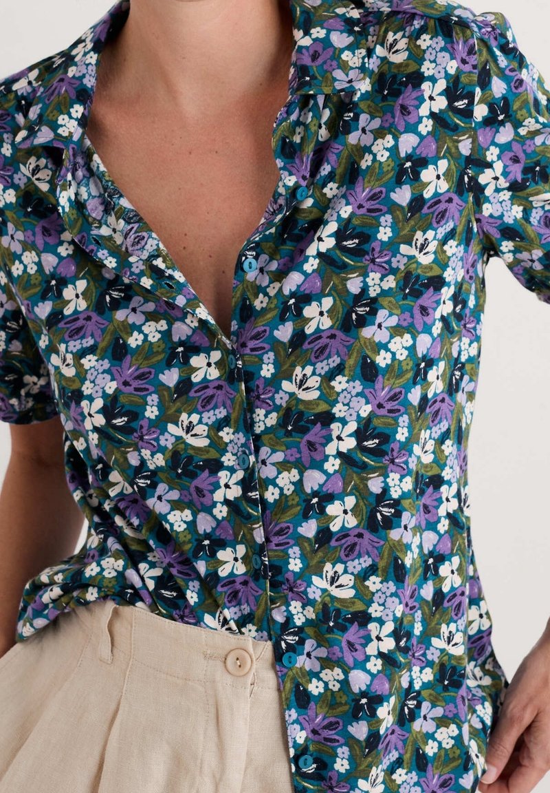 Gros plan d'une personne portant une chemise à manches courtes boutonnée avec des fleurs violettes, blanches et vertes et un pantalon beige.