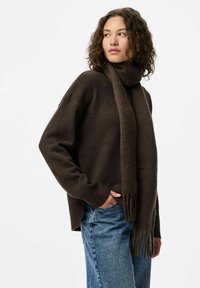 Ung kvinde med krøllet hår iført en mørkebrun sweater, matchende tørklæde og blå jeans, stående med den ene hånd i lommen og ser til siden.