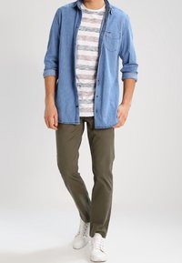 Chemise en denim bleu clair sur un T-shirt rayé blanc et gris, associée à un pantalon vert olive et des baskets blanches.