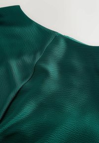 Tissu texturé vert foncé avec couture visible et détail de piqûre sur une surface lisse.
