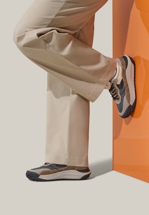 Sneaker grigio e marrone con materiali testurizzati, suola in gomma nera, lacci e branding discreto. Pantaloni beige parzialmente visibili.