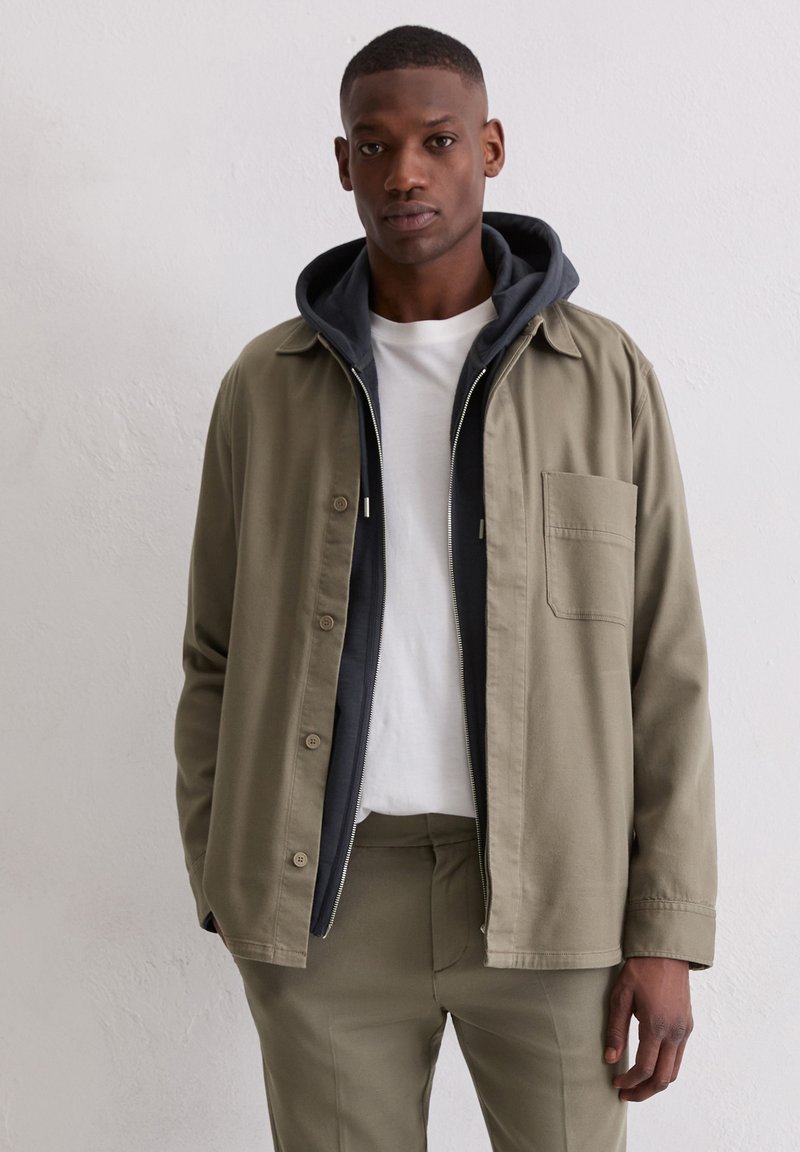 Olivgrüne Jacke mit Kragen, frontaler Tasche und Knopfverschluss, darüber eine marineblaue Kapuzenjacke und ein weißes T-Shirt. Schlanke Hose vervollständigt den Look.