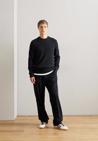 Man standing against plain wall wearing black sweater, black trousers, white sneakers with black stripes, hands in pockets, on wooden floor.