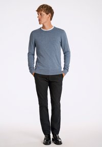 Maglione blu con tessuto strutturato, rifinitura del colletto bianca, abbinato a pantaloni neri e scarpe nere lucide, in posa in piedi su uno sfondo semplice.