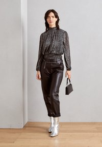 Blouse noire brillante à manches longues avec un col froncé, associée à un pantalon en cuir noir et des bottines métalliques argentées. Petite sac à main noir.