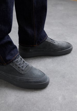 Graue Wildleder-Sneaker mit strukturiertem Obermaterial, flachen Gummisohlen und Schnürdesign, kombiniert mit dunklen Jeans aus Denim mit orangen Nähten.