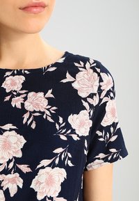 Abito floreale blu navy con maniche corte, che presenta fiori rosa chiaro e foglie verdi su un tessuto testurizzato. Collo rotondo e design aderente.
