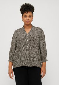 Blouse imprimé léopard en tissu léger ; dispose d'un col en V, d'une fermeture à boutons et de manches froncées avec des poignets élastiques. Les couleurs incluent le marron et la crème.
