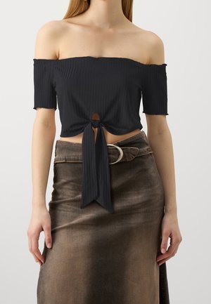 Femme portant un crop top noir épaules dénudées noué à la taille et une jupe marron taille haute avec une large ceinture et une boucle métallique.